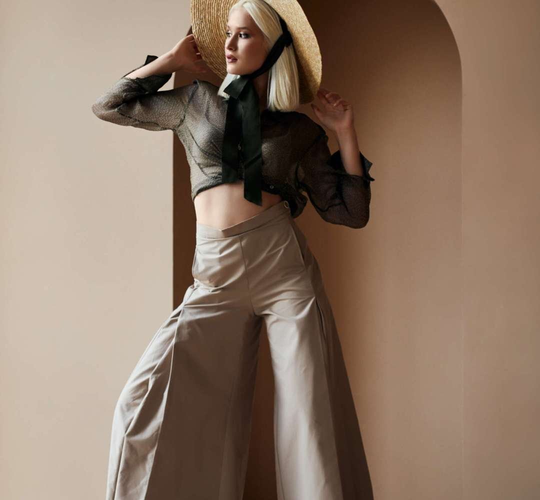 Attractive blonde woman wearing tied up blouse and wide beige trousers holding hand on her trendy straw and posing on camera. Young girl with bare torso and feet standing near arch in the wall